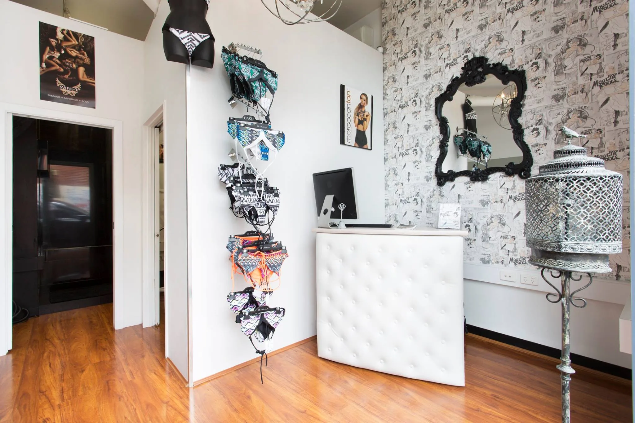 The salon reception — swimwear rack, tufted counter with iMac, baroque mirror on Moulin Rouge poster wall, crystal chandelier, and an open doorway to the private treatment room