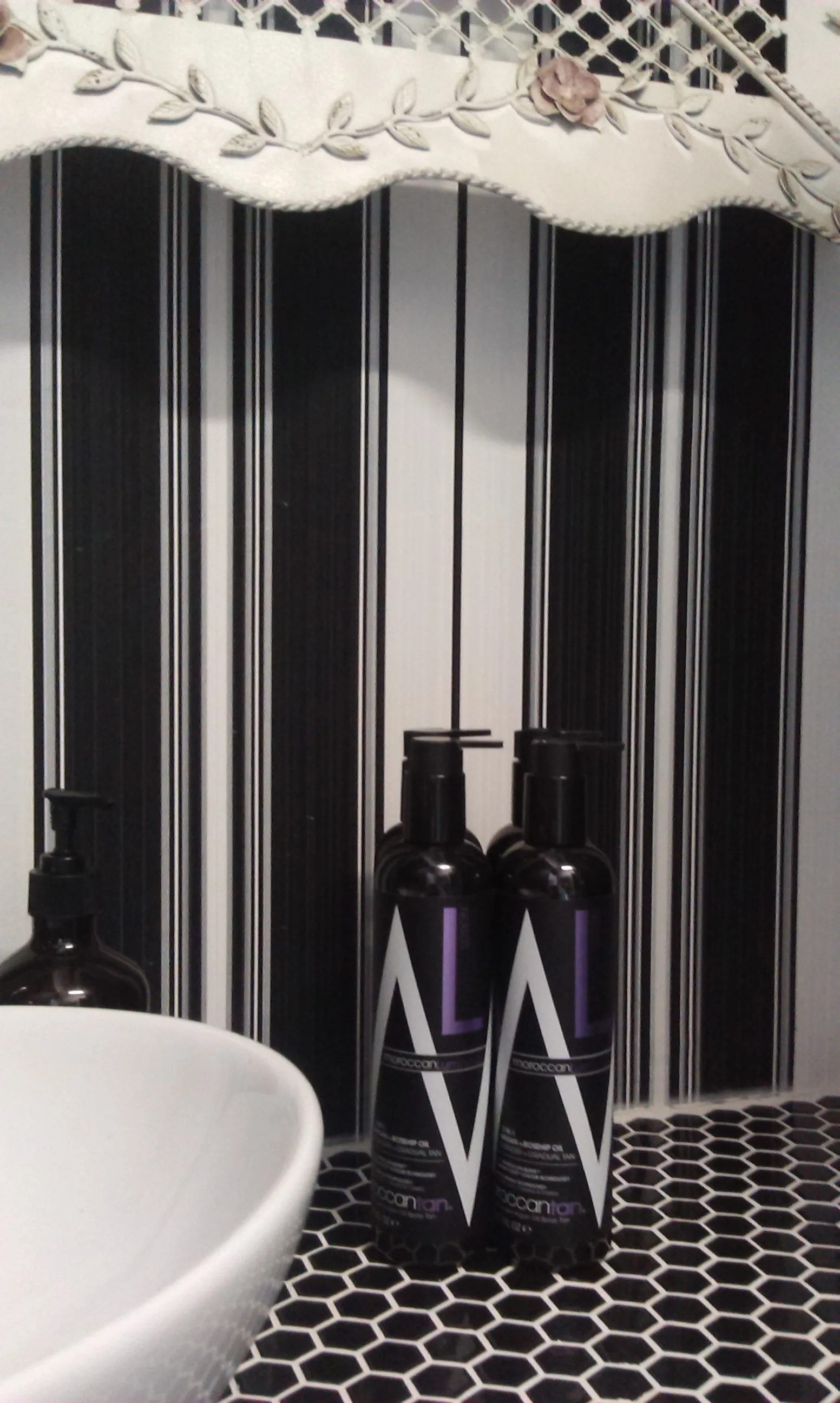 The private treatment room — black and white striped wallpaper, ceramic vessel basin, MoroccanTan pump bottles, ornate white mirror frame